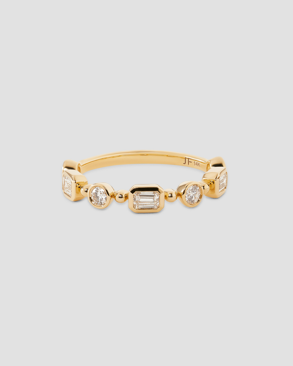 A gold band ring featuring a row of clear baguette and round-cut gemstones, displayed on a neutral light gray background.