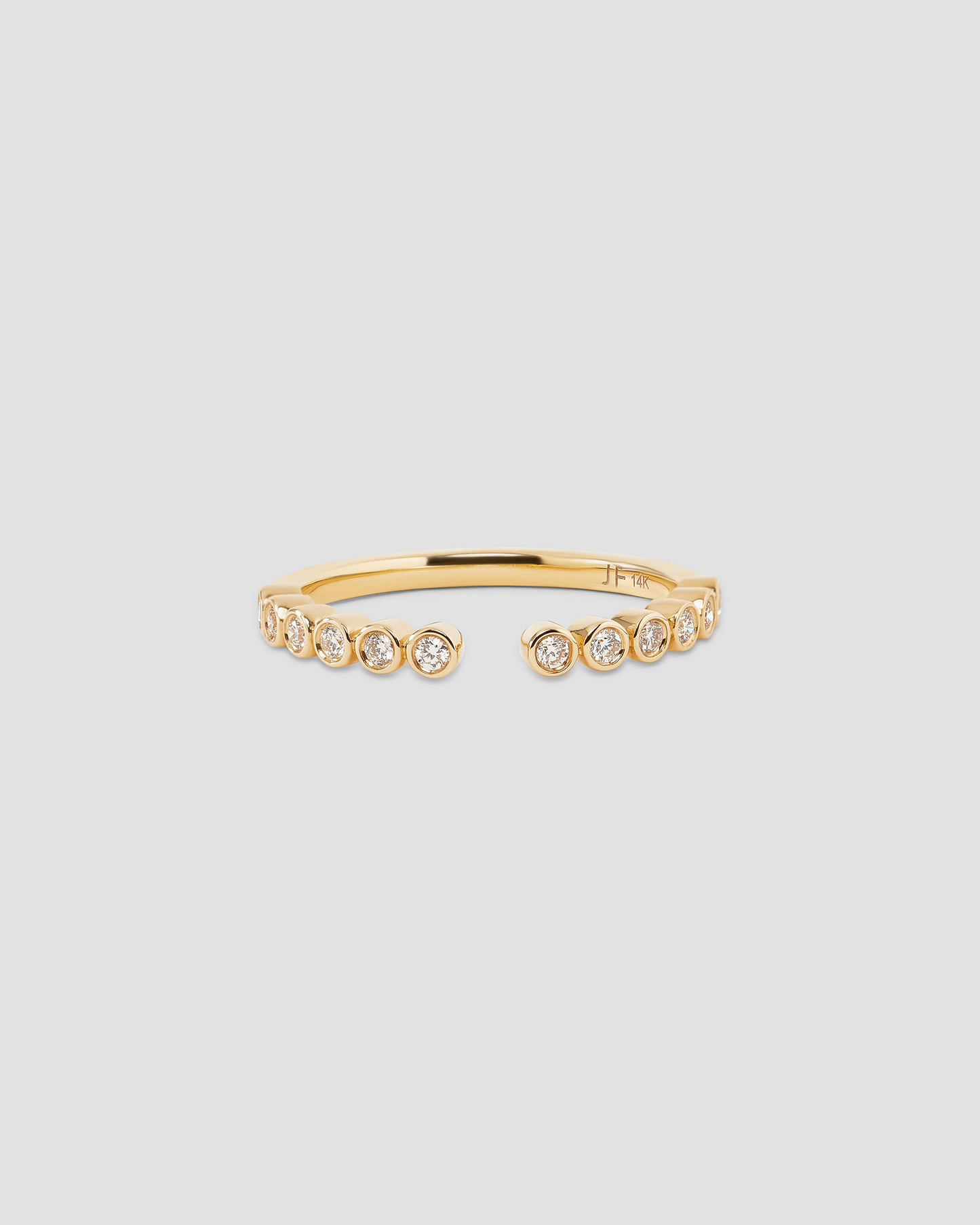 A delicate gold open ring featuring a row of small bezel-set round clear stones on the front, photographed against a solid light gray background.