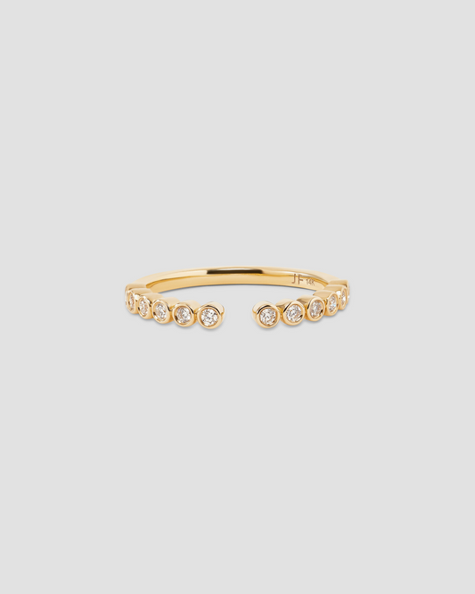 A delicate gold open ring featuring a row of small bezel-set round clear stones on the front, photographed against a solid light gray background.