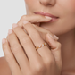 A close-up lifestyle shot of a model's hands resting near her lips, wearing a delicate rose gold ring with small clear gemstones on one finger, set against a soft, light gray background.