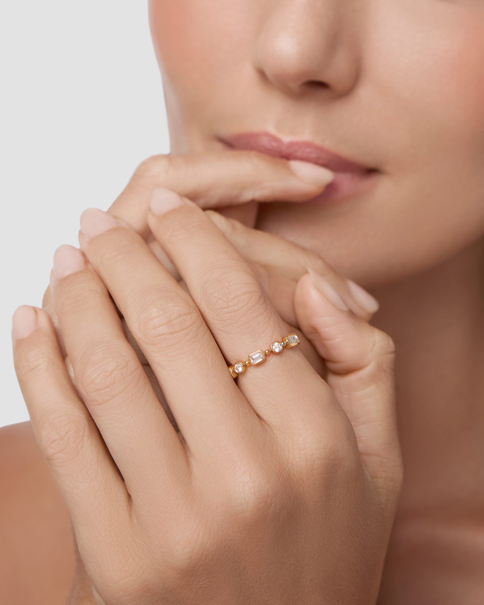 A close-up lifestyle shot of a model's hands resting near her lips, wearing a delicate rose gold ring with small clear gemstones on one finger, set against a soft, light gray background.